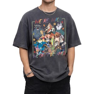 May include: A charcoal gray short-sleeve t-shirt featuring a colorful graphic print of various animated animal characters. The text "WE'RE WILD" appears at the top and "WE CAN'T BE TAMED" at the bottom, framed by a thin rectangular border.