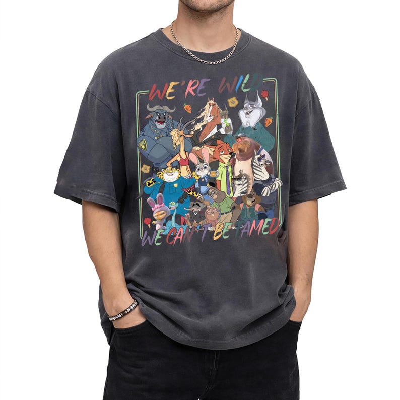 May include: A charcoal gray short-sleeve t-shirt featuring a colorful graphic print of various animated animal characters. The text "WE'RE WILD" appears at the top and "WE CAN'T BE TAMED" at the bottom, framed by a thin rectangular border.