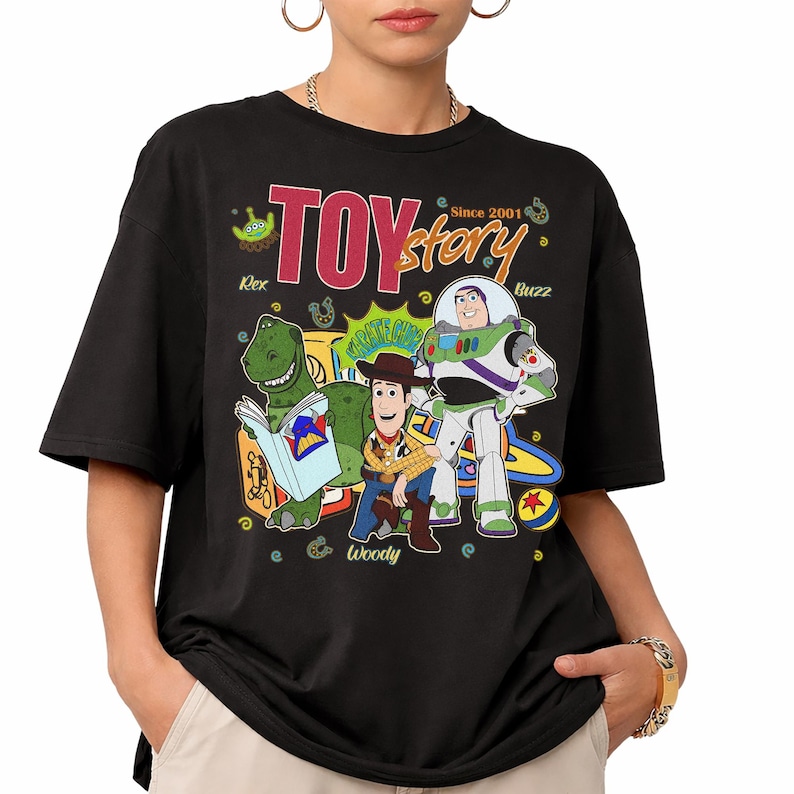 May include: Black t-shirt featuring a colorful graphic of Toy Story characters Rex, Woody, and Buzz Lightyear. The design includes the text "TOY STORY" and "Since 2001". The shirt has short sleeves and a crew neck.