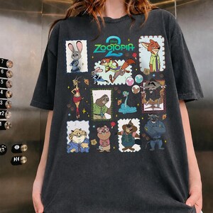 May include: A charcoal gray short-sleeve t-shirt featuring a graphic print of characters from the movie Zootopia 2. The design includes various animated figures in individual frames, including Judy Hopps, Nick Wilde, and others, with the Zootopia 2 logo.