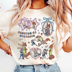 May include: An ivory t-shirt featuring a colorful graphic print of Zootopia characters including Judy Hopps and Nick Wilde. The design includes various illustrations of the characters, decorative frames, small flowers, and the text Welcome to Zootopia.