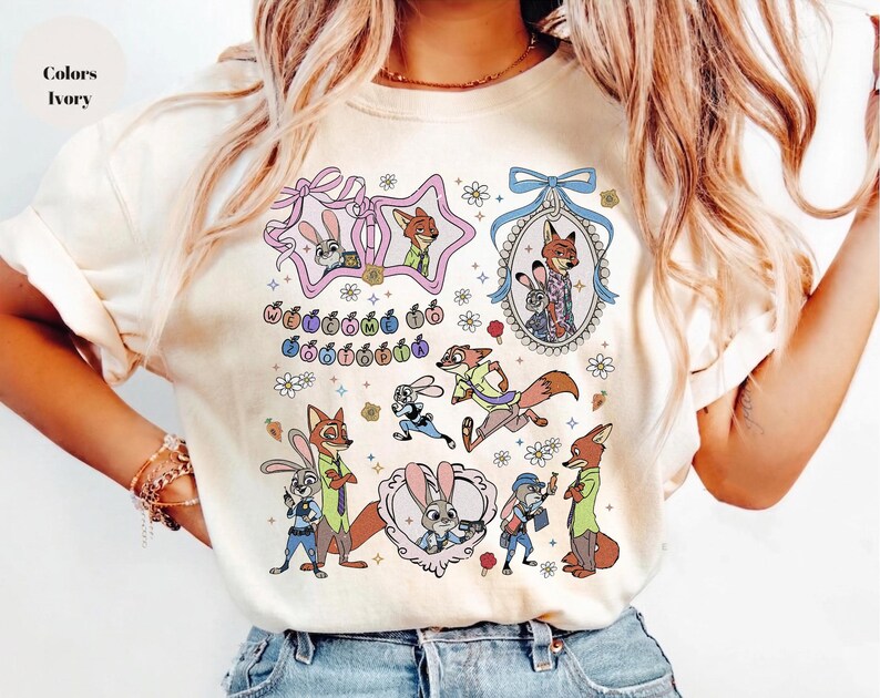 May include: An ivory t-shirt featuring a colorful graphic print of Zootopia characters including Judy Hopps and Nick Wilde. The design includes various illustrations of the characters, decorative frames, small flowers, and the text Welcome to Zootopia.