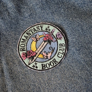 May include: Embroidered patch with the text "ROMANTASY BOOK CLUB" in a circular design. The patch features a sword, roses, butterflies, and a crescent moon in shades of pink, yellow, and blue, set against a denim background.