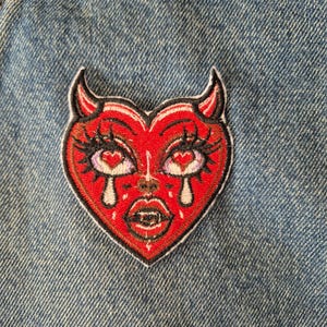 May include: A red heart-shaped patch with a devil face design. The patch features black horns, eyelashes, and a crying face with fangs. The patch is embroidered with a black border and is set against a blue denim background.