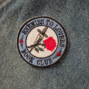 May include: Embroidered patch with the text "ENEMIES TO LOVERS BOOK CLUB" in a circular design. The patch features a dagger piercing a rose, with additional rose details. The patch is on a denim background.