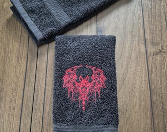 May include: Two dark gray hand towels with a red bat design embroidered on them. The towels are folded and have a textured appearance. The bat design is detailed and stylized, with a gothic theme.