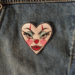 May include: Heart-shaped embroidered patch with a clown face design. The patch features a white background with a red heart-shaped nose, pink cheeks, and red lips. The eyes are outlined in black with teal eyeshadow and red accents.