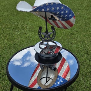 May include: A white cowboy hat featuring a brim adorned with blue and red rhinestones arranged in a star and stripe pattern. The hat rests on a black metal stand shaped like a horseshoe with a central star, positioned on a circular mirror surface.