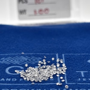 May include: A collection of small, sparkling diamonds scattered on a blue surface. The diamonds are clear and reflect light, creating a brilliant display. The background includes text and logos, with the words "GROWN DIAMONDS" visible.
