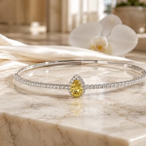 Puede incluir: Una pulsera rígida de tono plateado con una piedra preciosa amarilla central en forma de pera, rodeada por un halo de pequeñas piedras claras. La banda está incrustada con una fila continua de diminutas piedras claras, creando un aspecto brillante.