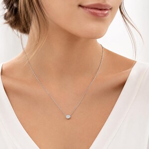 Puede incluir: Un collar de cadena de tono plateado con una piedra redonda, transparente y facetada, engastada en un bisel circular. El collar descansa sobre el cuello, mostrando una delicada cadena de eslabones metálicos unida al colgante de piedra central.