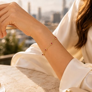 May include: A delicate gold chain bracelet featuring evenly spaced, round, vibrant red gemstones set in gold bezels. The bracelet rests on a wrist, showcasing a minimalist design with a thin, polished metal chain connecting each individual stone setting.