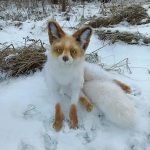 May include: A soft toy fox with a white body, brown paws, and a fluffy white tail. The fox has a white face with brown and tan markings and large, upright ears. It is sitting in the snow.