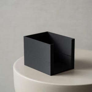 May include: A black, rectangular storage box with a textured surface. The box is open at the top and sits on a cream-colored, rounded surface. The background is a neutral gray.