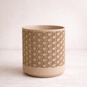 Puede incluir: Maceta cilíndrica beige con un diseño geométrico de estrellas. La maceta tiene una base ancha y un borde liso. El patrón de estrellas cubre la mayor parte de la superficie de la maceta. Aproximadamente 15 cm de altura.