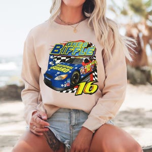 May include: Beige sweatshirt featuring a graphic of a race car with the text "Greg Biffle" and "National Guard" in blue, yellow, and white. The number "16" is prominently displayed in yellow. The design includes a checkered flag pattern.