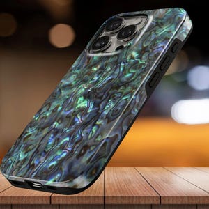 May include: A smartphone case with a pearlescent, iridescent design in shades of green, blue, and gray. The case has a black border and a camera cutout. The case is resting on a wooden surface.