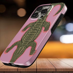 Könnte beinhalten: Eine Smartphone-Hülle mit einer detaillierten Illustration eines grünen Alligators mit texturierten Schuppen auf einem einfarbigen rosa Hintergrund. Die Hülle hat einen schwarzen Rahmen und Aussparungen für die Kameralinsen und Seitentasten.