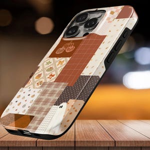 Könnte beinhalten: Eine Smartphone-Hülle mit einem Patchwork-Design aus herbstlichen Mustern, darunter Kürbisse, Geister, Karos und Schachbrettmuster in Braun-, Creme- und Orangetönen. Die Hülle hat einen schwarzen Rand und Aussparungen für die Kameralinsen.