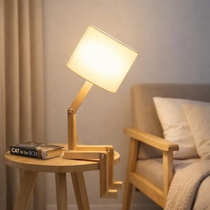May include: A wooden desk lamp with a square lampshade, positioned on a small round table. The lamp has a unique design with adjustable joints. A book titled "CAT in the BOX" rests on the table. The lamp is made of light-colored wood.