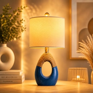 May include: A decorative table lamp with a blue and rope-textured base and a beige lampshade. The lamp has a gold-colored stem and a small, clear, spherical finial. The lamp is on a wooden surface.