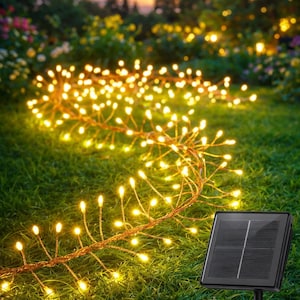 May include: Solar-powered string lights with warm white LED bulbs are arranged on a green lawn. The lights are connected by a brown wire and are near a black solar panel. The background features a sunset and trees.