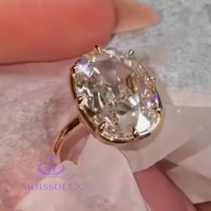 May include: A gold ring with a large, oval-cut, clear gemstone. The ring is held on a white cloth. The ring's band is thin and gold. The gemstone is set in a gold bezel setting. The ring is likely an engagement or fashion ring.