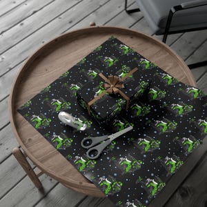 May include: A black wrapping paper featuring a repeating pattern of green snowmobiles and small white snowflakes. A gift box wrapped in the same paper is tied with a brown ribbon, accompanied by a clear tape dispenser and grey metal scissors.