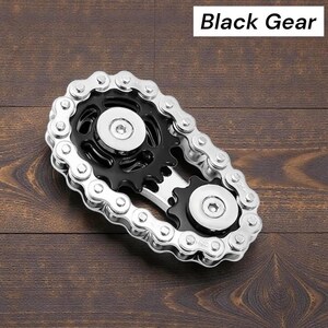 May include: A metal gear-shaped object with a black center and silver chain links. The text "Black Gear" is displayed in a white rectangle above the object. The object is on a dark brown wooden surface.