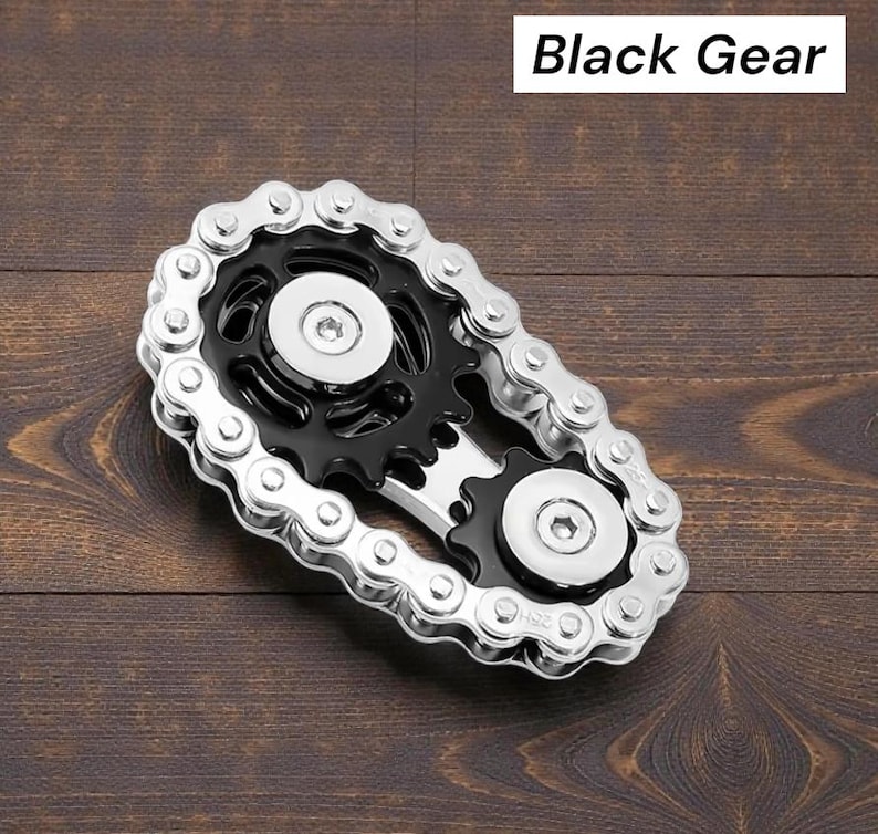 May include: A metal gear-shaped object with a black center and silver chain links. The text "Black Gear" is displayed in a white rectangle above the object. The object is on a dark brown wooden surface.