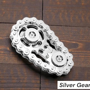 May include: A silver gear-shaped fidget toy with a chain design. The toy features two circular gears connected by a chain, with the text "Silver Gear" in a white box at the bottom. The object is on a wooden surface.