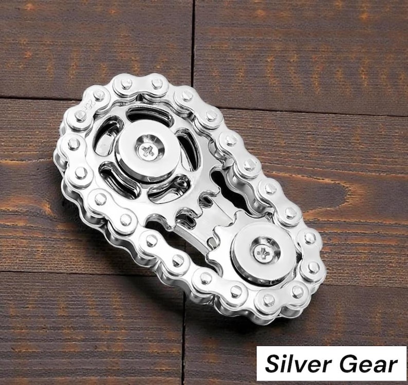 May include: A silver gear-shaped fidget toy with a chain design. The toy features two circular gears connected by a chain, with the text "Silver Gear" in a white box at the bottom. The object is on a wooden surface.