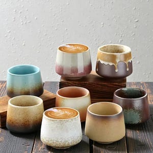 May include: A collection of handcrafted ceramic coffee cups in various colors, including blue, brown, and beige. Some cups are filled with coffee, showcasing latte art. The cups are arranged on a wooden surface, creating a rustic aesthetic.