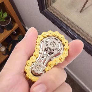 May include: A gold and silver fidget spinner in the shape of a bicycle chain and gears. The spinner is held in a person's hand, showcasing its intricate design and metallic finish. The gold chain surrounds a silver gear mechanism.