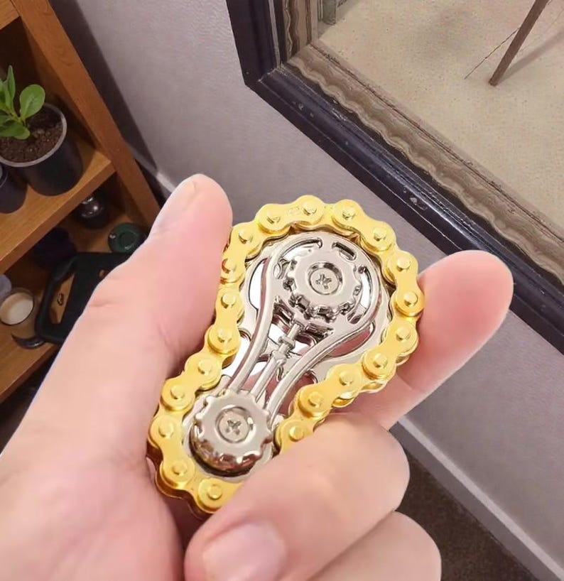 May include: A gold and silver fidget spinner in the shape of a bicycle chain and gears. The spinner is held in a person's hand, showcasing its intricate design and metallic finish. The gold chain surrounds a silver gear mechanism.