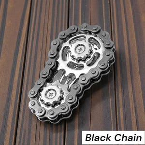 May include: A silver and black metal chain design with gears and the text "Black Chain" on a wooden surface. The chain is arranged in an oval shape, with two gears in the center.