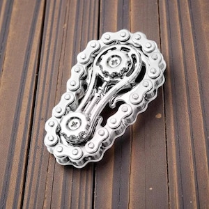 May include: A silver-colored metal belt buckle with a motorcycle chain design. The buckle features a gear-like center and is set against a wooden background. The chain links are detailed and create a unique accessory.
