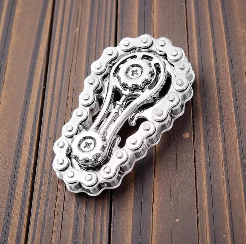 May include: A silver-colored metal belt buckle with a motorcycle chain design. The buckle features a gear-like center and is set against a wooden background. The chain links are detailed and create a unique accessory.