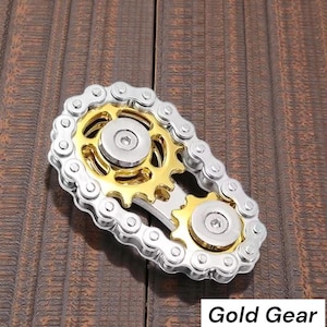 May include: A silver and gold metal fidget toy with a gear and chain design. The toy has a gold center with silver gears and a chain that surrounds the gears. The text "Gold Gear" is in the bottom right corner.