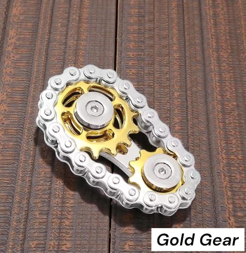 May include: A silver and gold metal fidget toy with a gear and chain design. The toy has a gold center with silver gears and a chain that surrounds the gears. The text "Gold Gear" is in the bottom right corner.