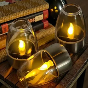 May include: Three battery-operated candle lanterns with a warm, flickering light. Each lantern has a clear glass shade and a silver-coloured base. The lanterns are arranged on a wooden surface, creating a cosy ambiance.