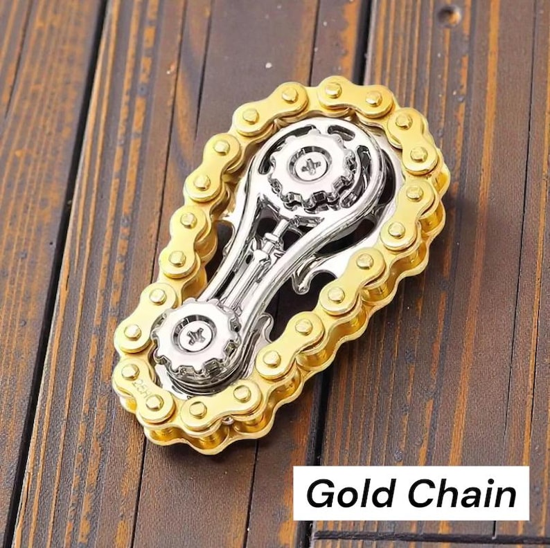 May include: A gold-colored chain surrounds a silver-colored metal object with gears. The object is on a wooden surface. The text "Gold Chain" is in a white rectangle at the bottom.