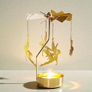 May include: A gold-colored candle holder with a spinning top and hanging fairy ornaments. The candle holder has a round base with a lit candle inside. The fairies are suspended from the top and rotate when the candle is lit.