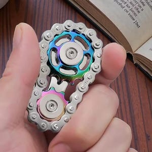 May include: A silver metal fidget spinner in the shape of a bicycle chain and gears. The central gear has a rainbow iridescent finish, with shades of blue, green, and purple. The spinner is held in a person's hand.