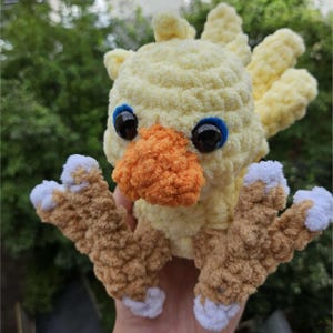 May include: A handmade, plush yellow bird with an orange beak, blue-rimmed black eyes, and brown and white feet. The bird is made of soft, textured yarn and has a cheerful, cartoonish appearance. The bird is a handmade item.