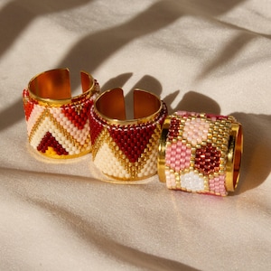 May include: Three gold-toned rings with beaded designs in red, white, and pink. The rings have a wide band and a split design. The beads are arranged in geometric patterns. The rings are displayed on a cream-colored fabric.
