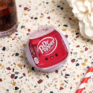 May include: A round, clear plastic container featuring a red Dr. Pepper soda can graphic on the lid. The graphic includes the white Dr. Pepper logo, a small smiling face with two black eyes and a mouth, and nutritional text printed on the side.