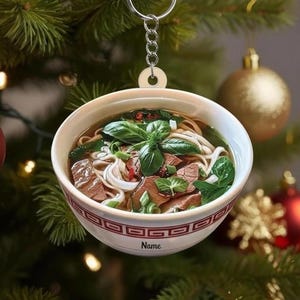 May include: A round, bowl-shaped ornament featuring a detailed illustration of a noodle soup with beef, green herbs, and broth. The exterior of the bowl has a red geometric pattern and the word Name printed on the side. It hangs from a silver keychain.