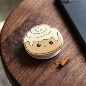May include: A circular phone grip featuring a light brown cinnamon roll illustration with white icing, dark brown eyes, a small smile, and pink cheeks. The accessory has a white base and is designed to attach to the back of a mobile device.