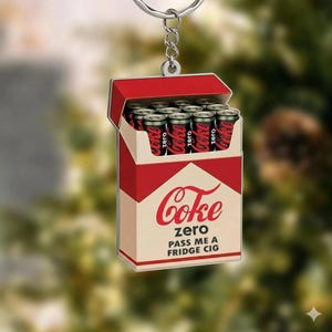 May include: A novelty keychain featuring a red and white box of Coke Zero cans. The box has the Coke Zero logo and the text "PASS ME A FRIDGE CIG". The keychain is attached to a silver chain.